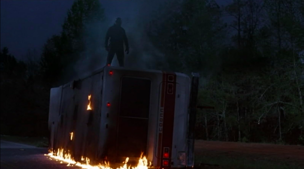 Jason est debout sur un camping-car renversé, qui commence à prendre feu